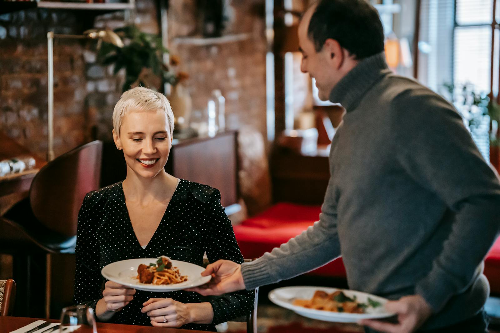 Positive adult couple having dinner in bright room near table while man serving plates with tasty pasta with meatballs near chairs in daylight