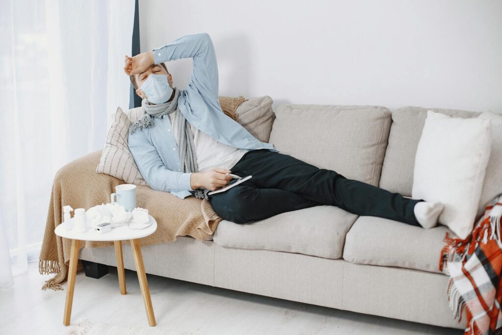 Sick man with mask and scarf resting on couch at home, feeling unwell.
