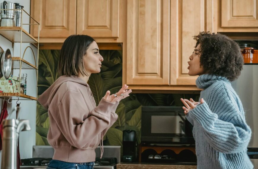 Side view of unhappy young multiracial girlfriends in casual clothes standing in kitchen while having disagreement and looking at each other