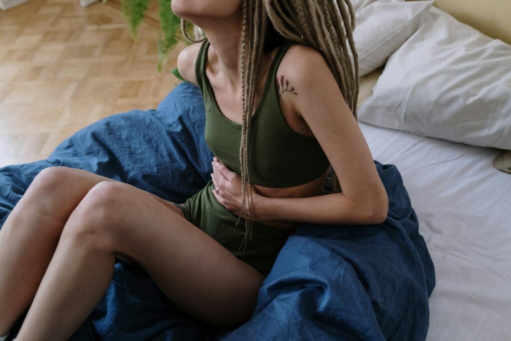 Woman sitting in bed holding her stomach, indicating pain or discomfort, possibly due to cramps.