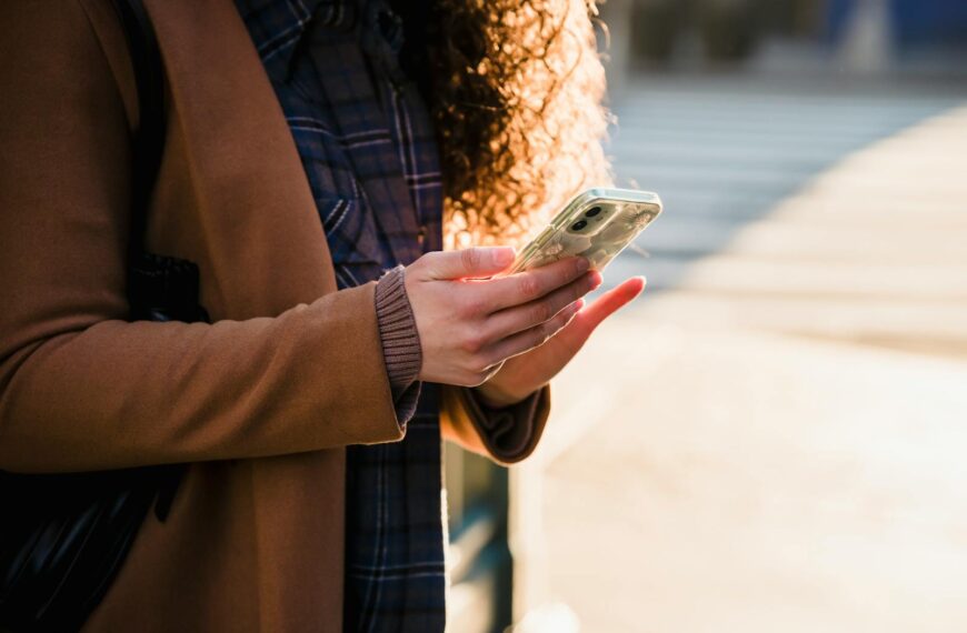 Crop anonymous female with curly hair in warm stylish coat using mobile phone on blurred background of urban street