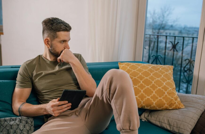 Bearded man sitting on a sofa, thoughtfully holding a tablet indoors.