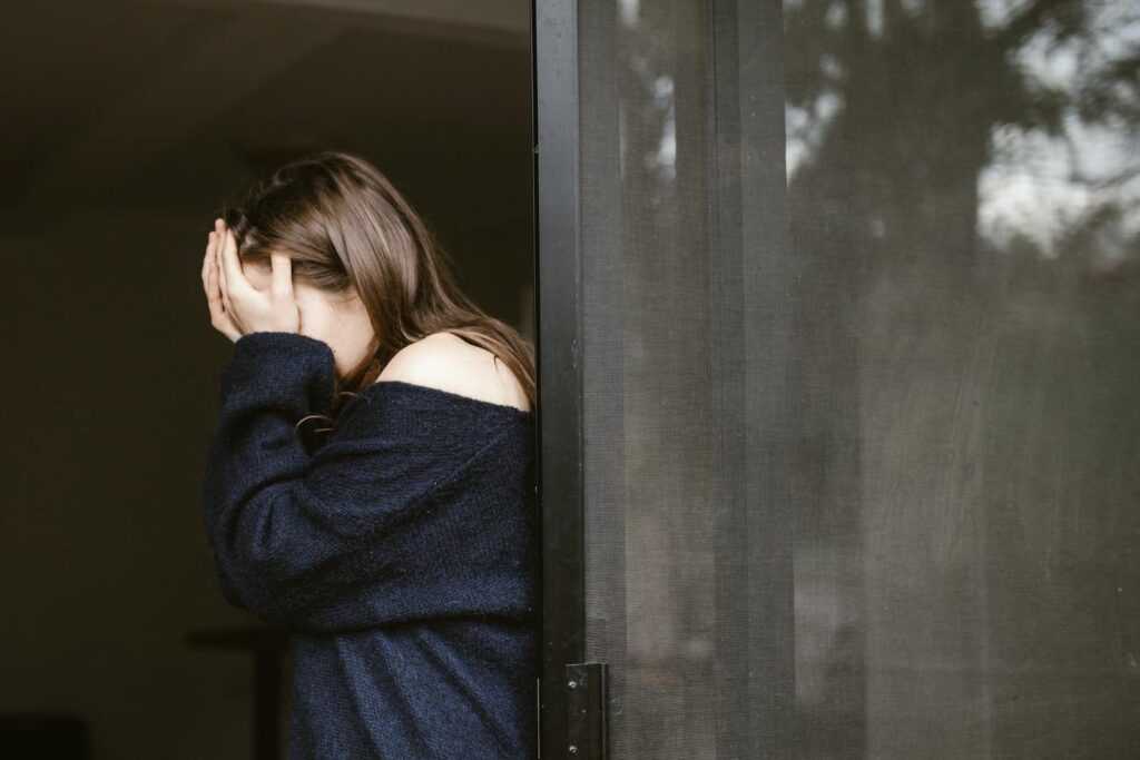 A woman stands indoors by a sliding door, covering her face with both hands.