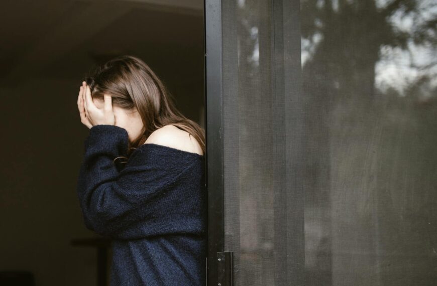 A woman stands indoors by a sliding door, covering her face with both hands.
