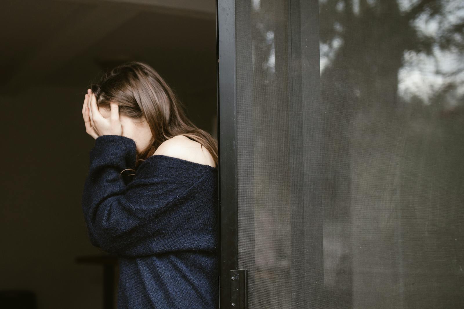 A woman stands indoors by a sliding door, covering her face with both hands.