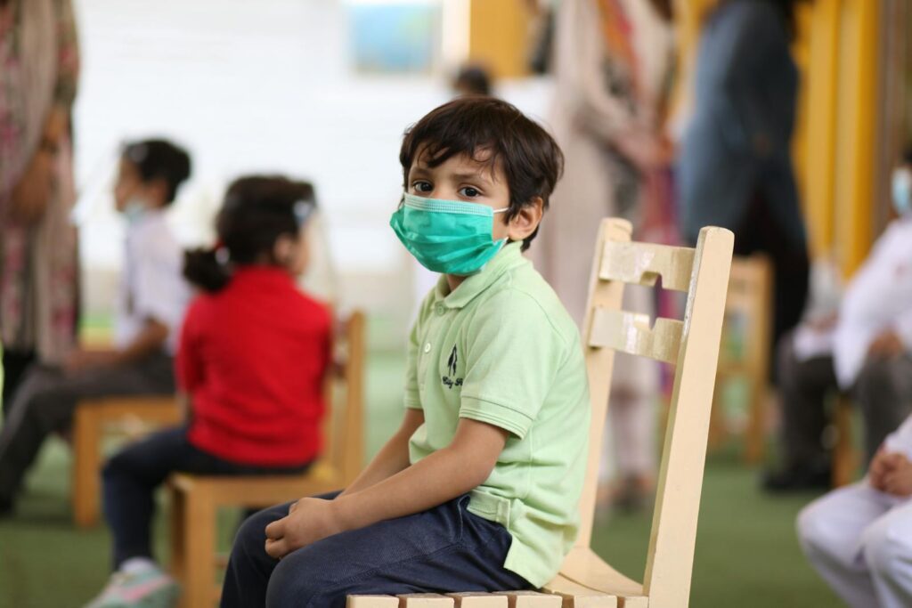 A young child wearing a mask sits on a chair in a classroom, emphasizing education during COVID-19.