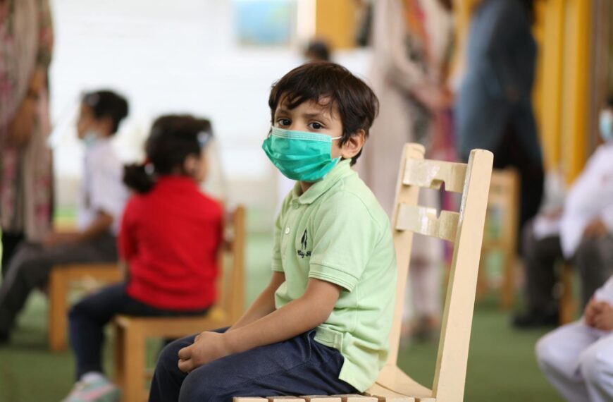 A young child wearing a mask sits on a chair in a classroom, emphasizing education during COVID-19.