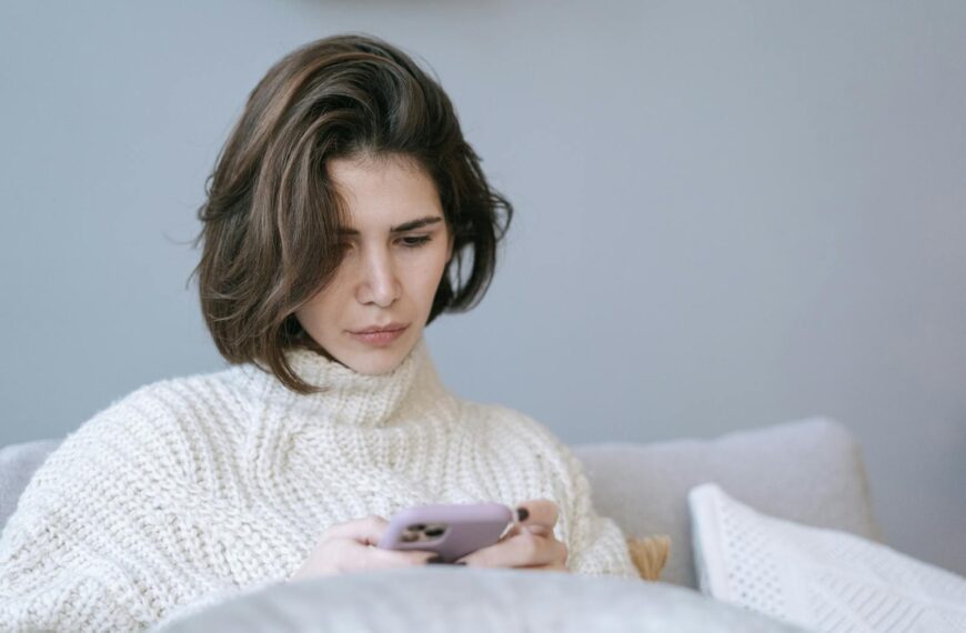 Woman in cozy knit sweater using a smartphone indoors, expressing a thoughtful mood.
