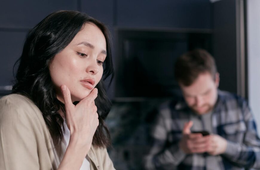Young woman pondering indoors while man uses smartphone in the background.