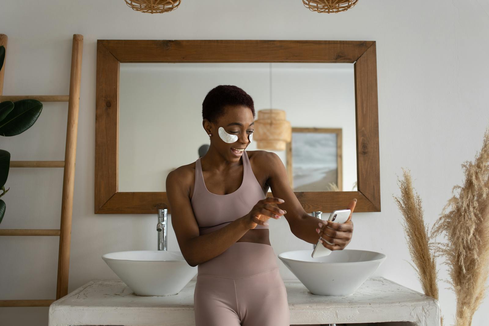 Young woman smiling in a bathroom while using skincare patches and video calling on phone.