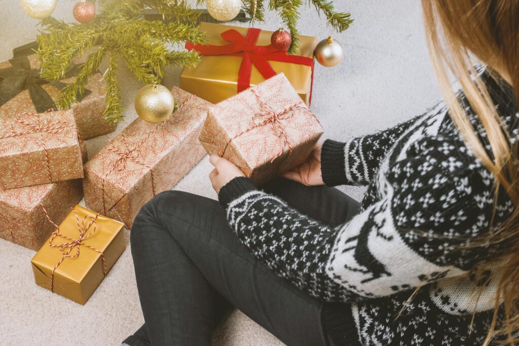 Woman in festive sweater holding gift by a decorated Christmas tree.