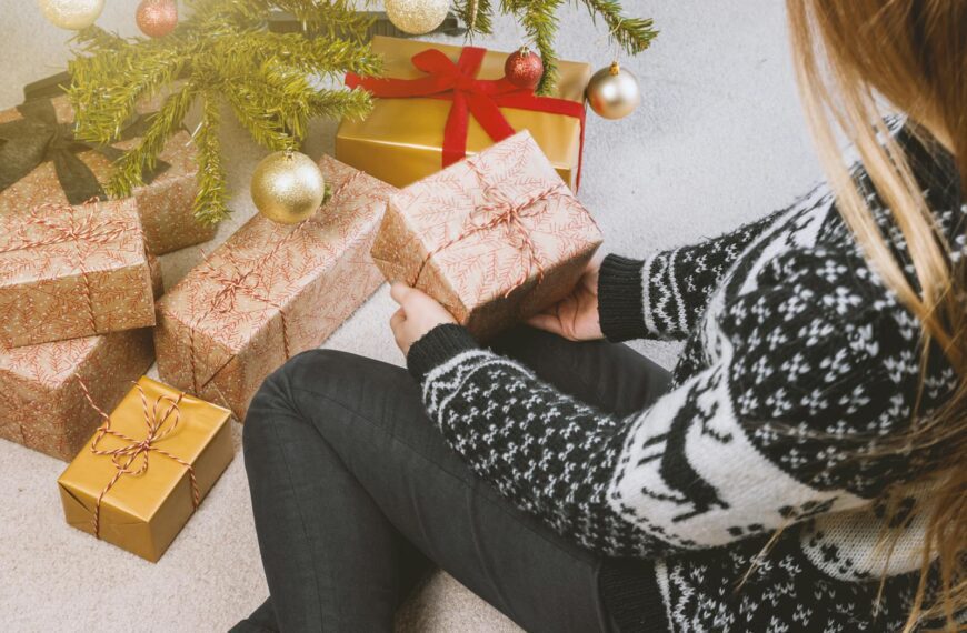 Woman in festive sweater holding gift by a decorated Christmas tree.