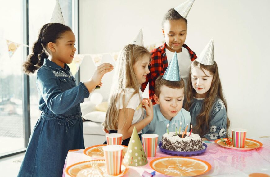 A joyful group of kids celebrating a birthday indoors with cake and party hats.