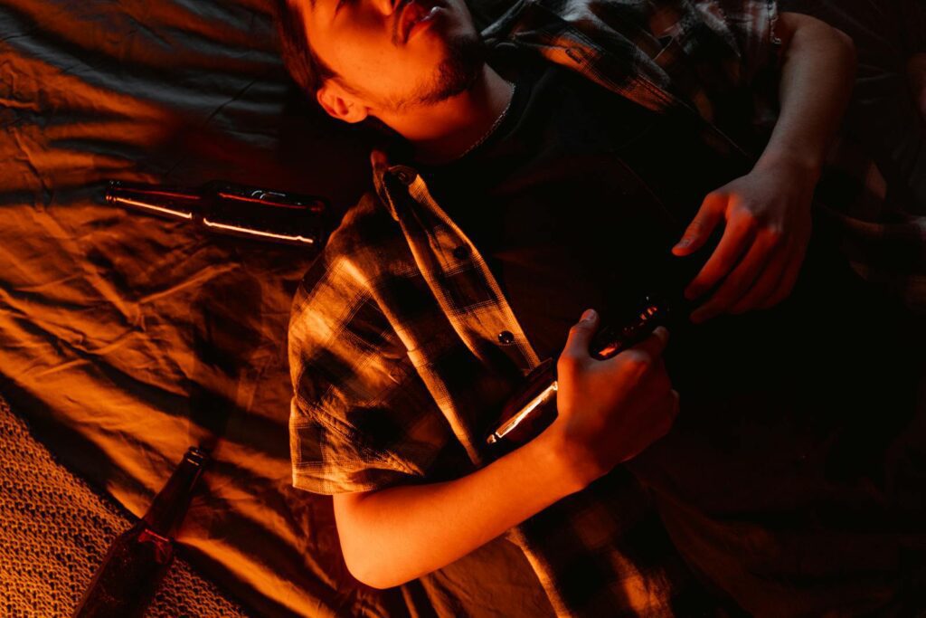 A young adult male lies on a blanket in orange light, holding bottles.
