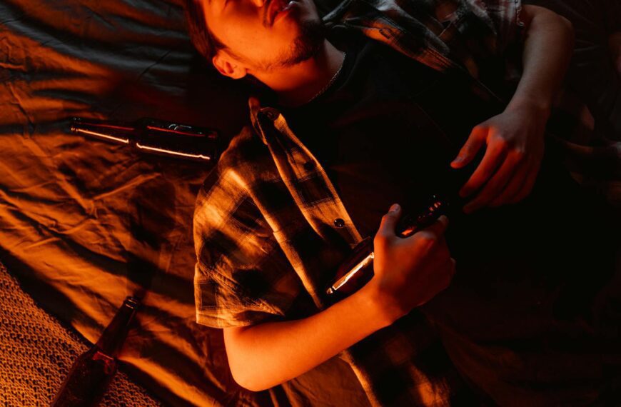 A young adult male lies on a blanket in orange light, holding bottles.