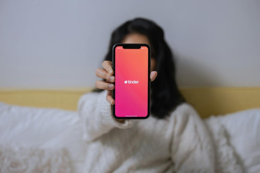 A woman in casual attire displays the Tinder app on her smartphone while sitting indoors.