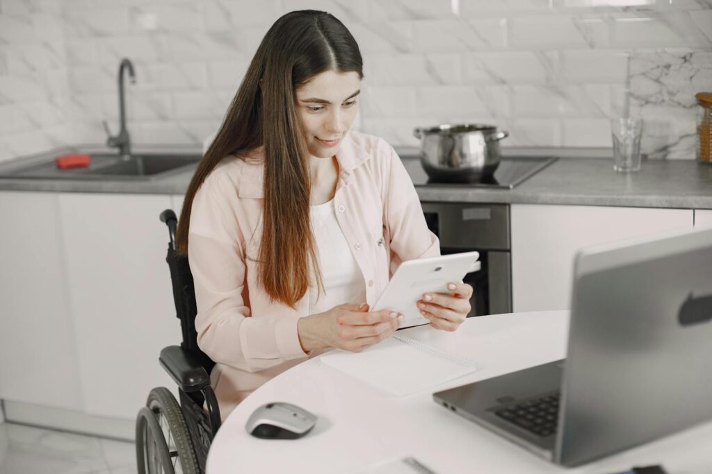 Woman in wheelchair engaged with tablet at kitchen table, creating an inclusive work environment.