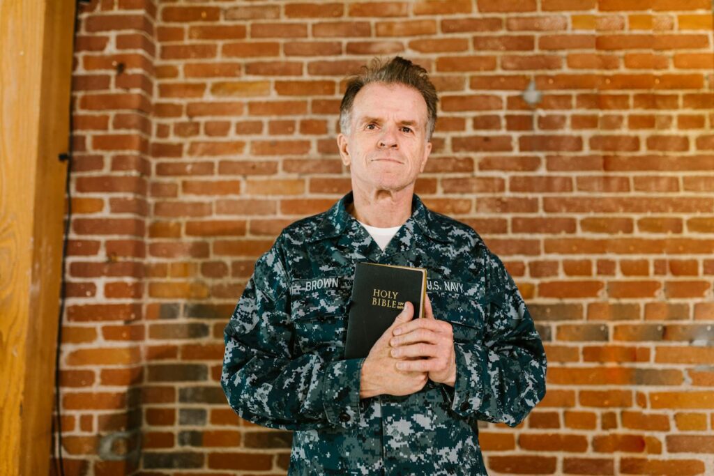 A senior U.S. Navy veteran holding a Holy Bible against a brick wall background.