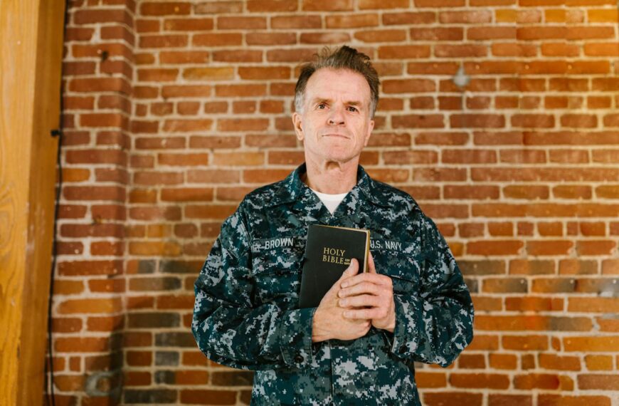 A senior U.S. Navy veteran holding a Holy Bible against a brick wall background.