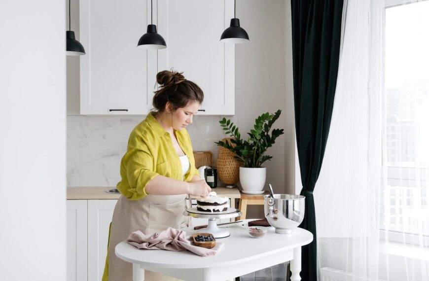 Concentrated housewife in apron preparing delicious cake with cream placed on stand on white table with ingredients in modern light kitchen