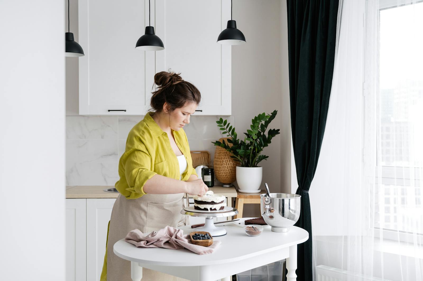 Concentrated housewife in apron preparing delicious cake with cream placed on stand on white table with ingredients in modern light kitchen