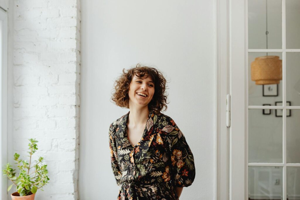 Happy woman in a floral top, standing indoors by a white wall and plant, smiling.