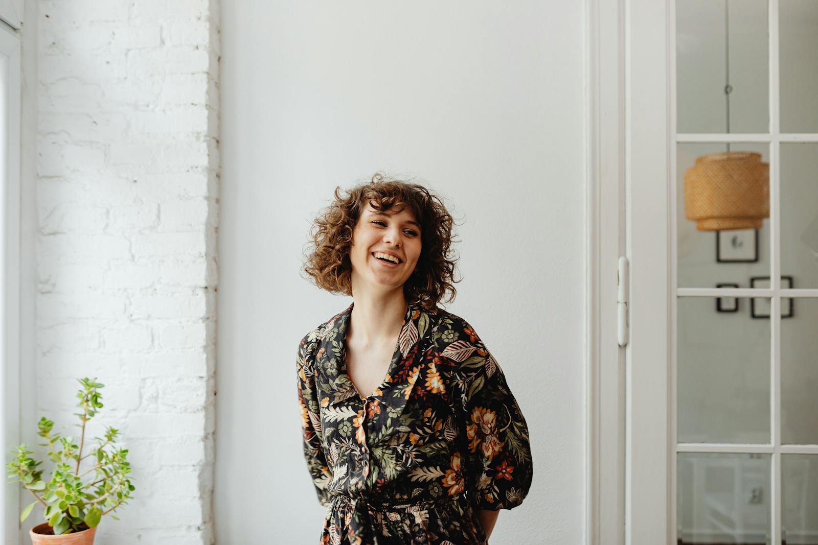 Happy woman in a floral top, standing indoors by a white wall and plant, smiling.