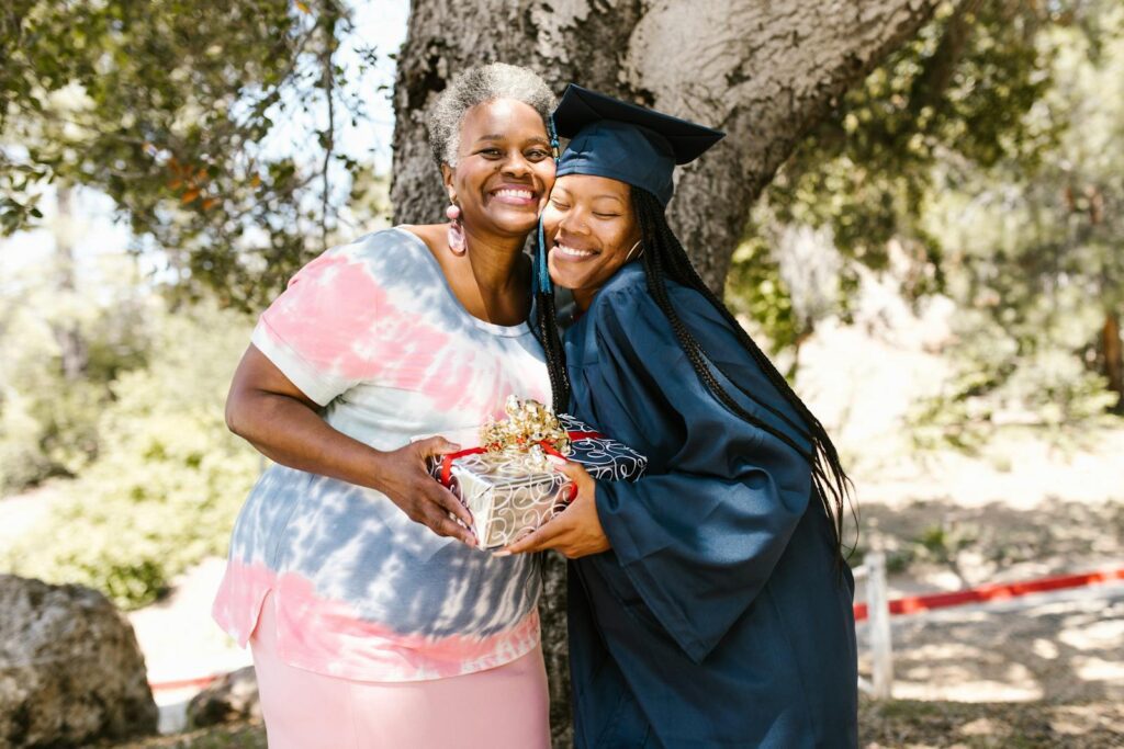 Graduate hugging her mother, celebrating with a gift under a tree.