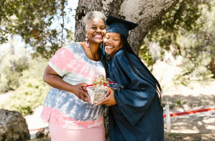Graduate hugging her mother, celebrating with a gift under a tree.