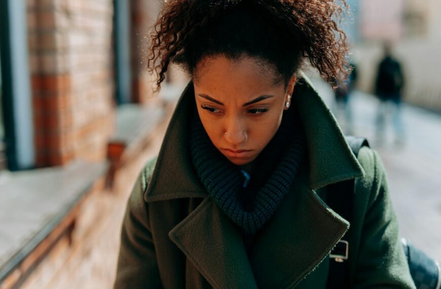 A young woman in a trench coat appears thoughtful and pensive walking down a city street.