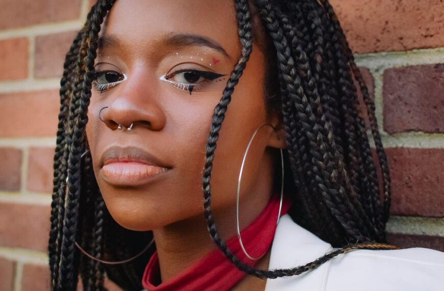 Close-up portrait of a woman with braided hair, hoop earrings, and nose piercing against a brick wall.