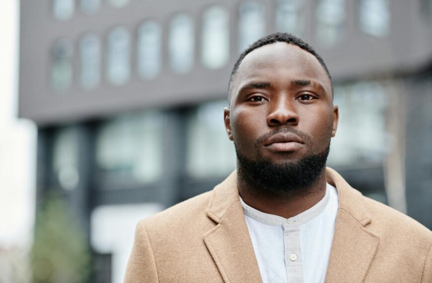 Portrait of a confident black man in a stylish coat, facing camera with urban backdrop.