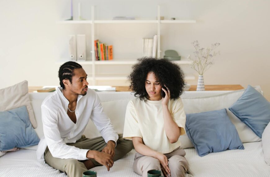 African American couple relaxing at home, sitting on sofa, talking and using phone.