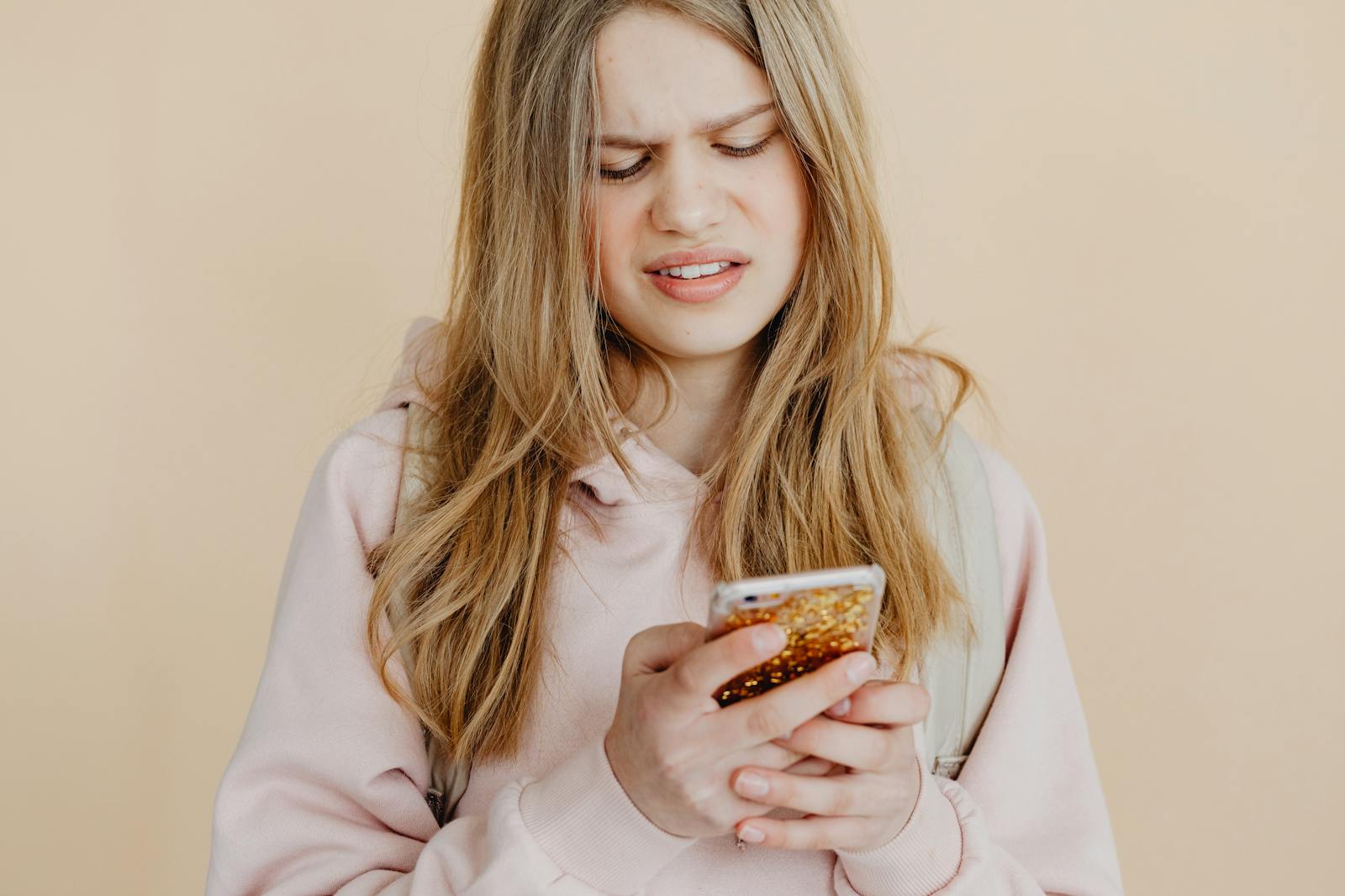 Teen girl with blonde hair in pink hoodie using smartphone, expressing curiosity indoors.