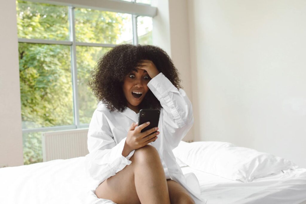 Amazed woman sitting on bed, expressing shock while using her smartphone.
