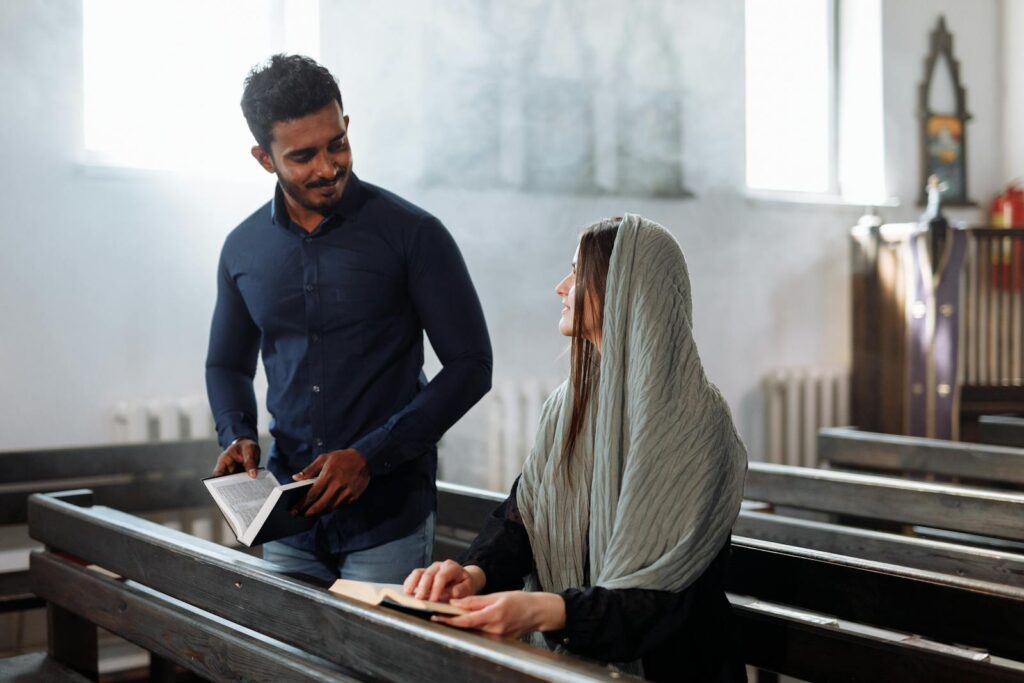 A South Asian man and woman seated in a church pew reading together, ambient light and peaceful setting.
