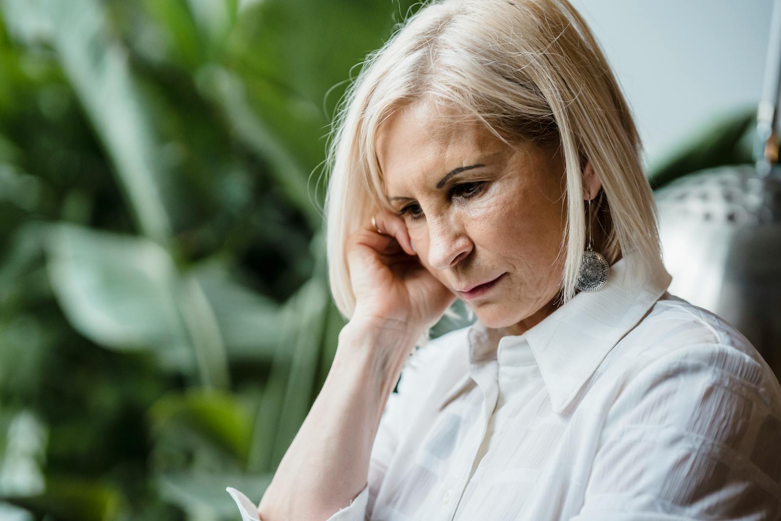 Senior woman in thoughtful pose surrounded by greenery indoors.