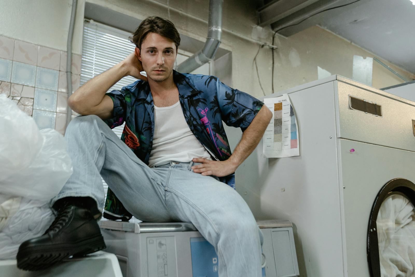 Man in casual attire poses confidently atop a washing machine in an industrial laundry room.