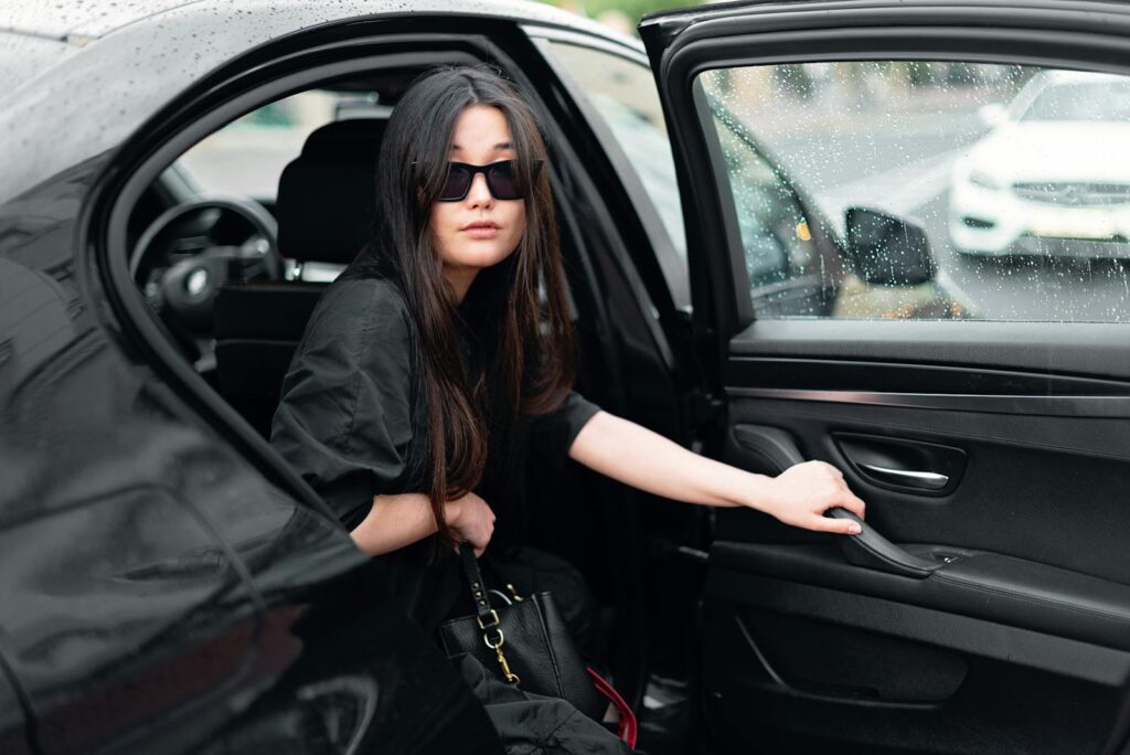 Stylish woman with sunglasses stepping out of a black luxury car in the rain.
