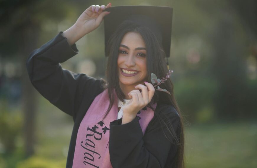 Young graduate woman smiling in cap and gown, celebrating her success outdoors.
