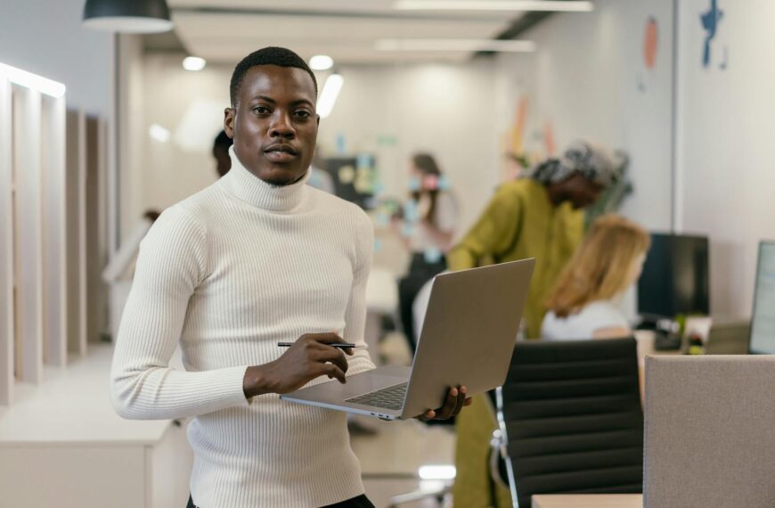 Confident man in white turtleneck working on laptop in a modern office setting.