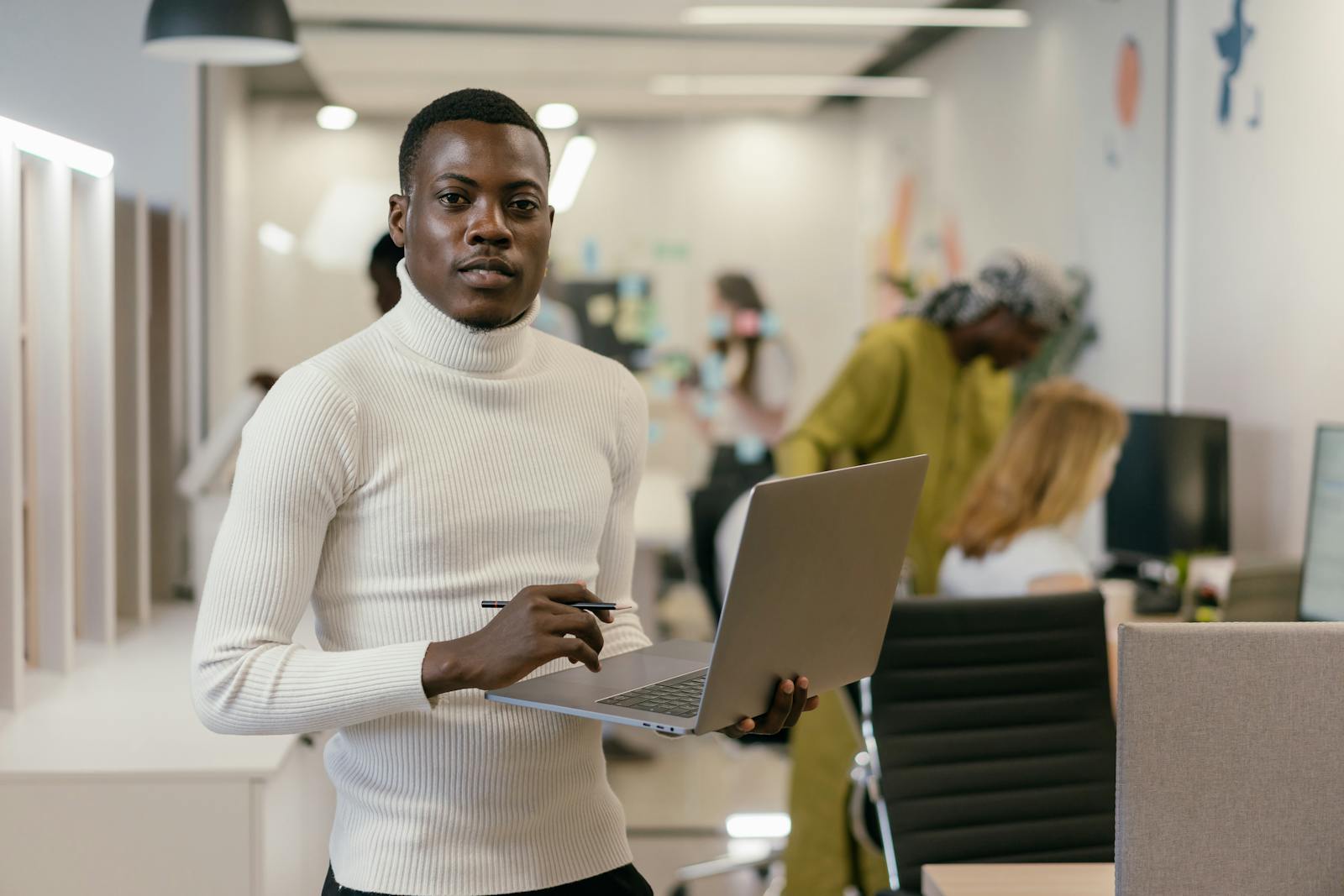 Confident man in white turtleneck working on laptop in a modern office setting.