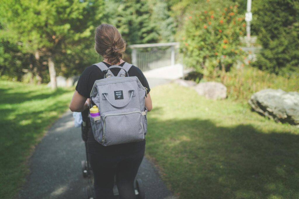 Woman strolling through a park with a backpack and stroller, enjoying a sunny day.