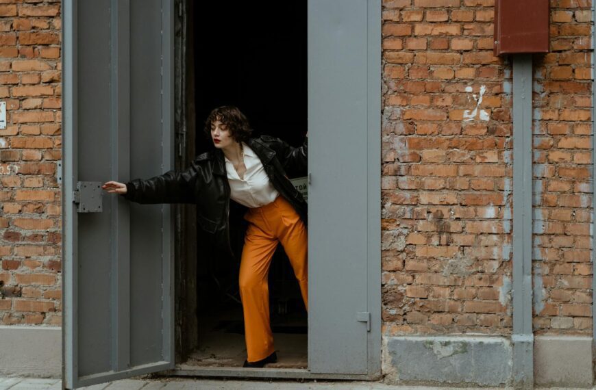 A woman in vibrant fashion opens a large industrial door set in a brick wall.