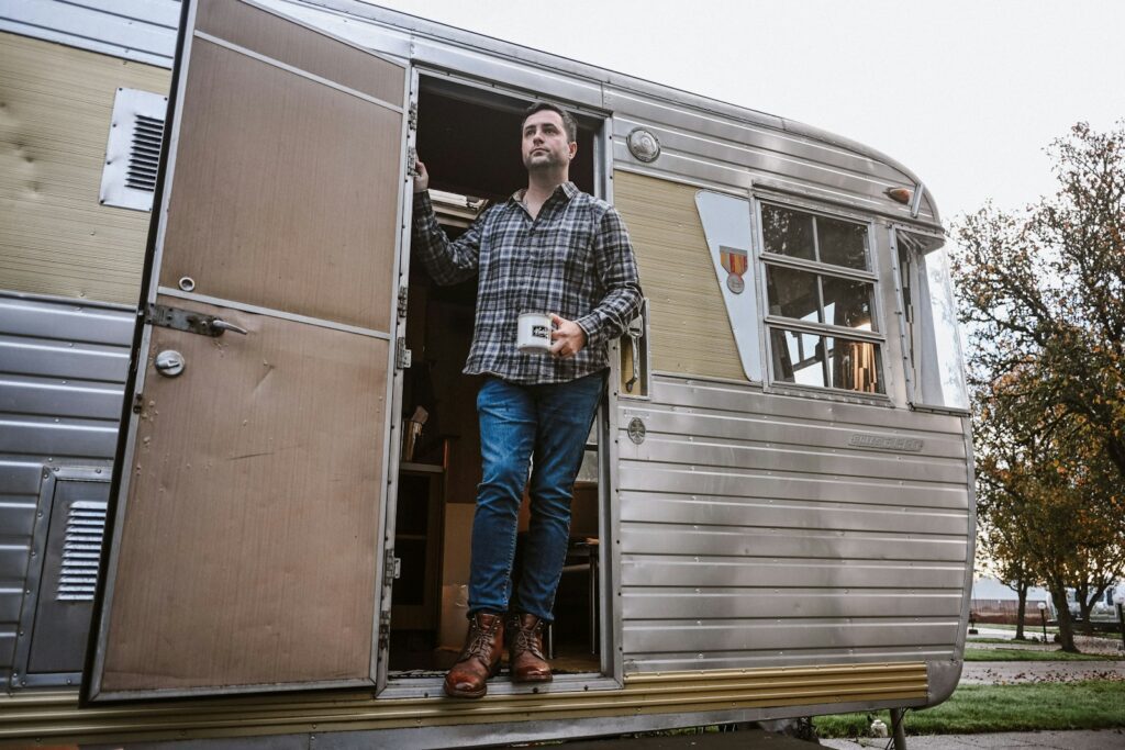 A man exits a vintage camper holding a coffee mug.