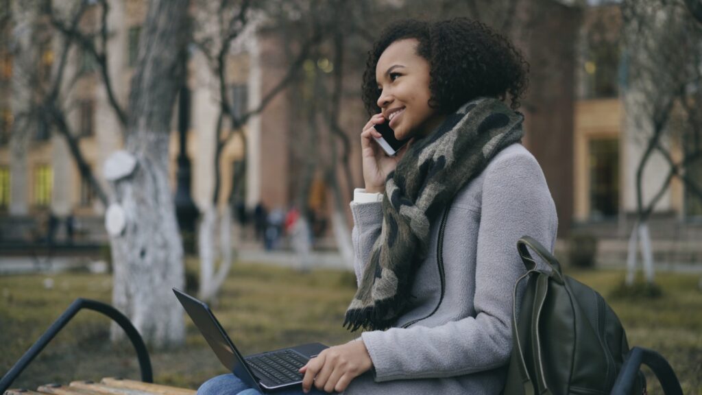 Young woman talking on phone with laptop outdoors