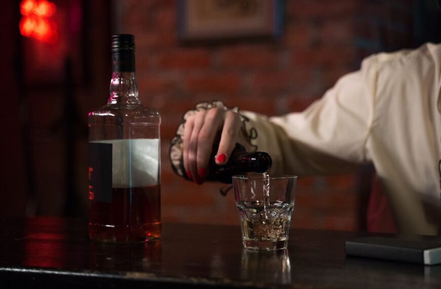 A hand with red nail polish pouring whiskey into a glass at a bar with a brick wall backdrop.