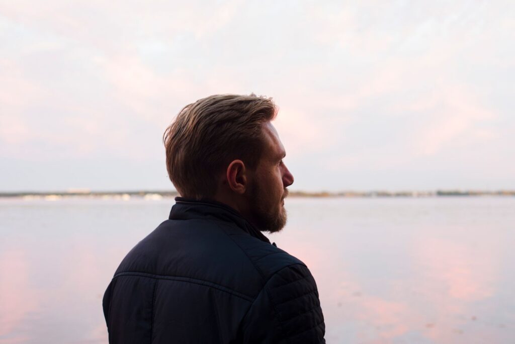 A serene side profile shot of a man gazing over a calm lake at sunset, capturing a moment of reflection.