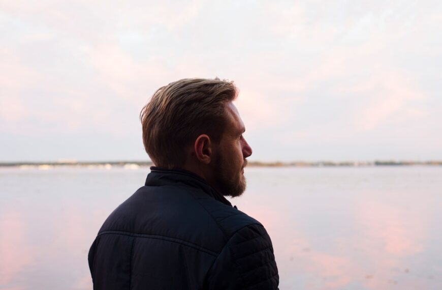 A serene side profile shot of a man gazing over a calm lake at sunset, capturing a moment of reflection.