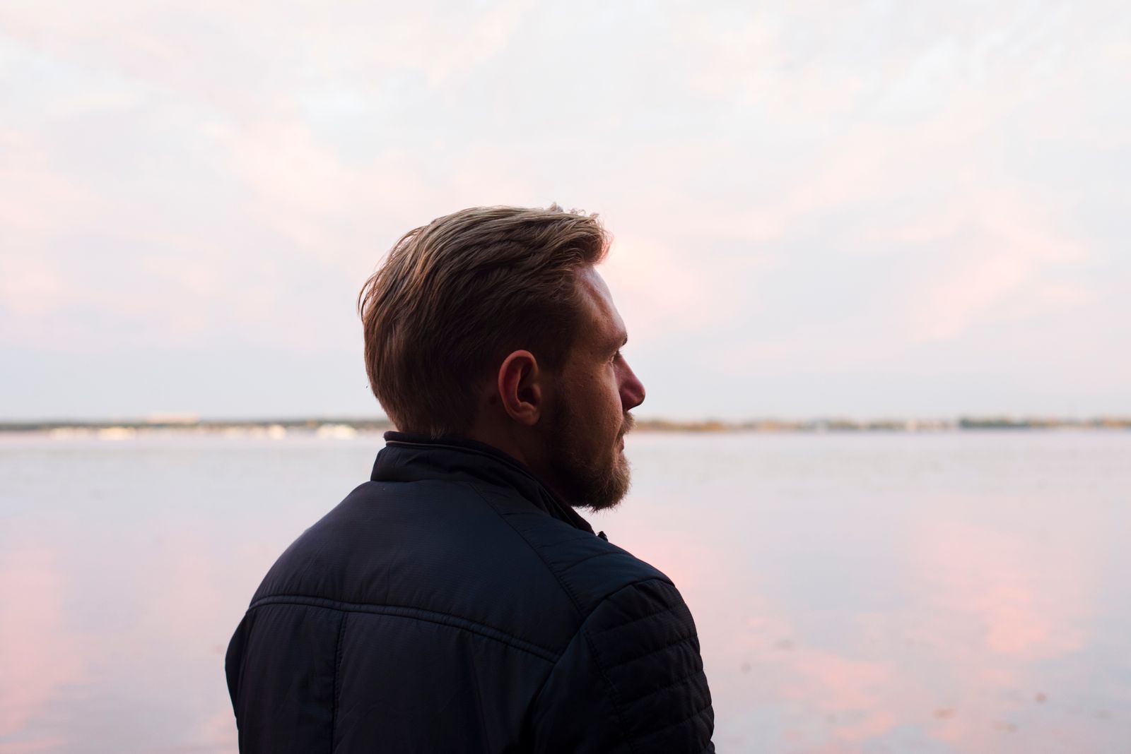 A serene side profile shot of a man gazing over a calm lake at sunset, capturing a moment of reflection.